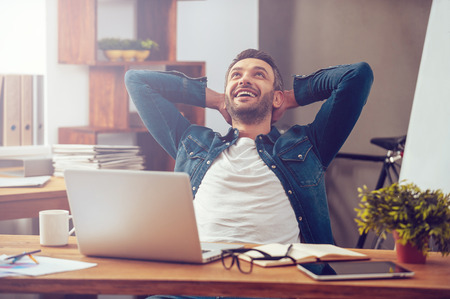 Satisfied with work done. Happy young man working on laptop while sitting at his working place in office Satisfied with work done. Happy young man working on laptop while sitting at his working place in office