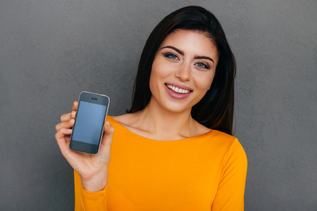 Look what I got! Attractive young woman showing her smart phone and smiling while standing against grey backgroundの写真素材