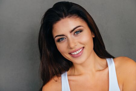 Portrait of beauty. Beautiful young woman in white tank top looking at camera and smiling while standing against grey backgroundの写真素材