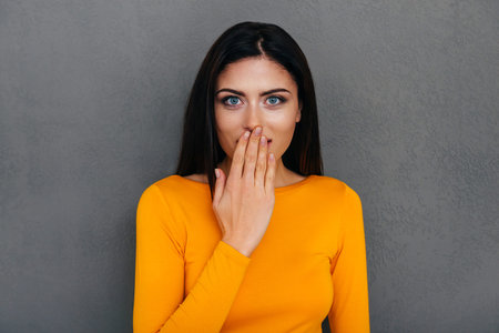 Outstanding news! Surprised young woman covering mouth with hand and looking at camera while...