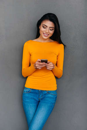Quick message to friend. Attractive young woman holding smart phone looking at it with smile while standing against grey backgroundの写真素材