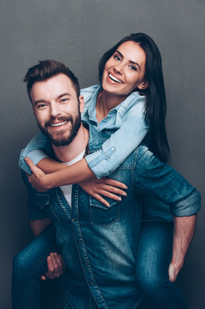 Having fun together. Handsome young man piggybacking beautiful woman and smiling while standing against grey backgroundの写真素材