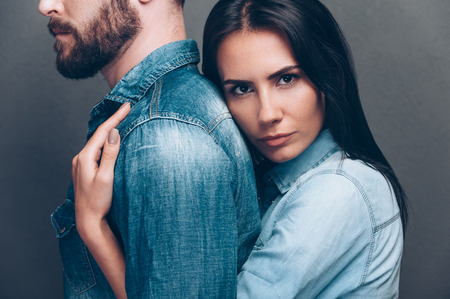 He is my everything. Side view of beautiful young woman bonding to back of beard man and looking at camera while both standing against grey backgroundの写真素材