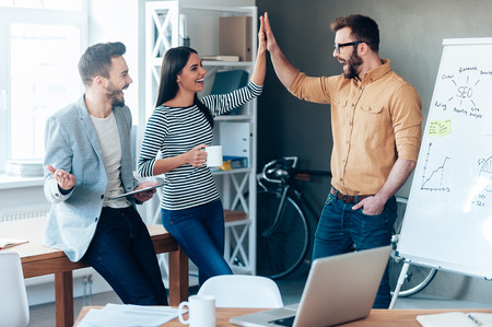 Celebrating success. Happy young man standing near whiteboard in office and giving high five to his colleagues Celebrating success. Happy young man standing near whiteboard in office and giving high five to his colleagues