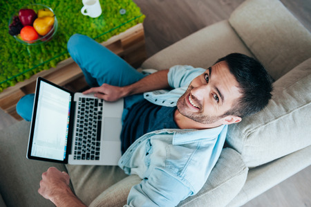 Enjoying carefree time at home. Top view of handsome young man working on laptop and smiling while sitting on the couch at homeの写真素材