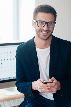 Smart and confident. Confident young man holding smart phone and smiling while leaning at the office deskの写真素材