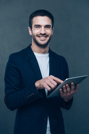 Enjoying his new digital tablet. Cheerful young man holding digital tablet and smiling while standing against grey backgroundの写真素材