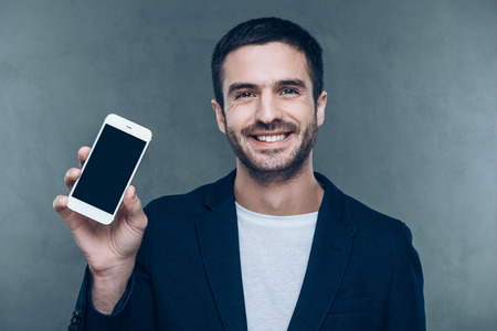Look at my new smart phone! Cheerful young man holding mobile phone and smiling while standing against grey backgroundの写真素材