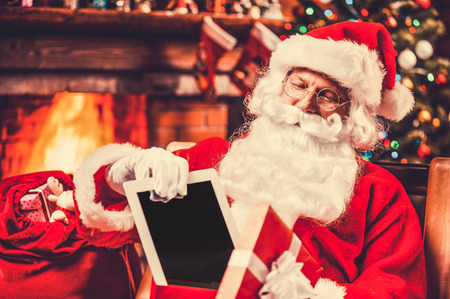 The best present! Cheerful Santa Claus putting a digital tablet into the gift box and smiling while sitting at his chair with Christmas Tree in the backgroundの写真素材