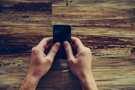 Typing quick message. Close-up top view of male hands holding mobile phone over the rustic wooden...