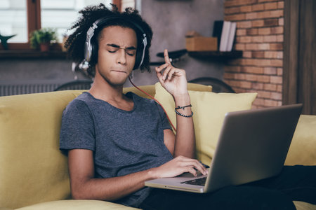 This playlist is great! Young African man working on laptop and wearing headphones while sitting on...