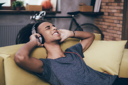 Enjoying music at home. Cheerful young African man wearing headphones while sitting on the couch at homeの写真素材