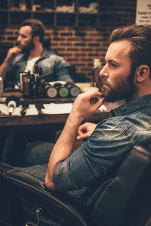 Looking awesome as ever. Side view of handsome young bearded man looking away and keeping hand on chin while sitting in chair at barbershopの写真素材