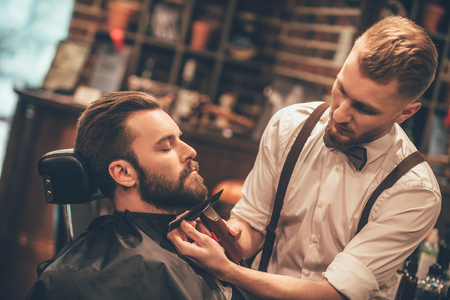 Grooming of real man. Side view of young bearded man getting beard haircut at hairdresser while sitting in chair at barbershopの写真素材