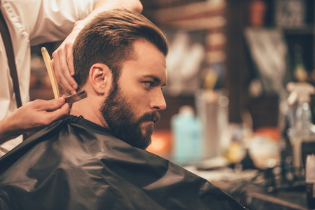 Making hair look magical. Close-up side view of young bearded man getting haircut with straight edge razor by hairdresser at barbershopの写真素材