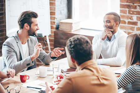 Great proposition. Group of five young people discussing something with smile while sitting at the office tableの写真素材