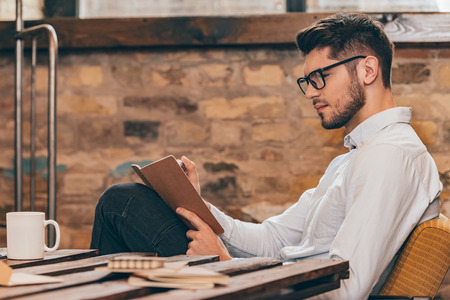 Noting his ideas. Side view of handsome young man in glasses writing in his note pad while sitting at his working placeの写真素材