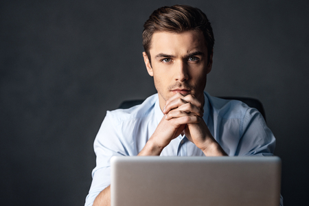 Concentration and elegance. Confident handsome young man looking at camera and keeping handsclasped while sitting with laptop against black backgroundの写真素材