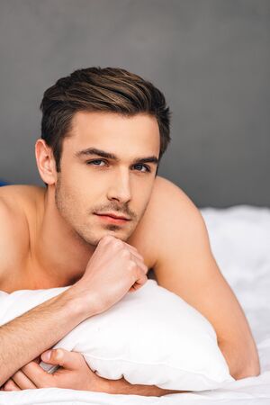 Day dreaming in bed. Pensive young man looking at camera and holding hand on chin while lying on front in bed against grey backgroundの写真素材