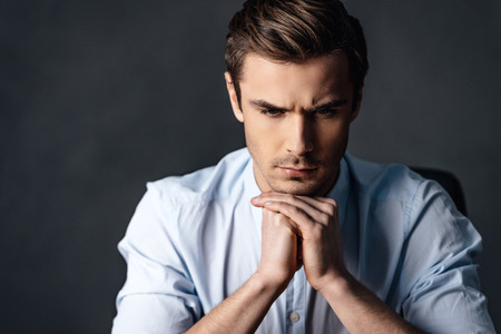 Calm and handsome. Portrait of handsome young man keeping hands clasped and looking thoughtful while sitting against black backgroundの写真素材