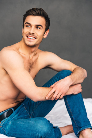 Feeling happy. Cheerful handsome young man looking away with smile while sitting on bed against grey backgroundの写真素材