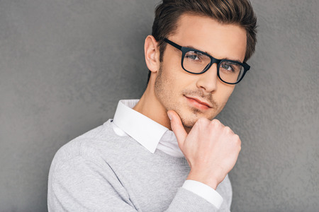 Confident and intelligent. Confident young man in glasses holding hand on chin and looking at camera while standing against grey backgroundの写真素材