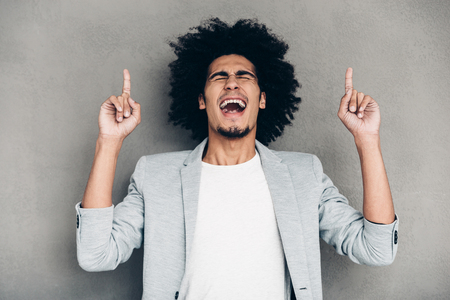 All the fun is there!Cheerful young African man pointing up and keeping mouth open while standing against grey backgroundの写真素材