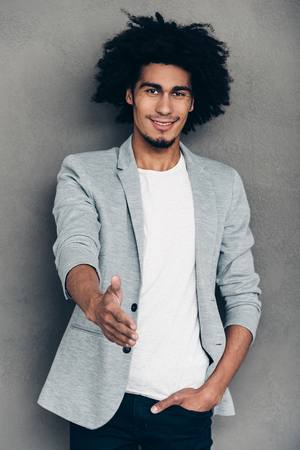 Nice to meet you! Cheerful young African man reaching out his hand in greeting you while standing against grey backgroundの写真素材