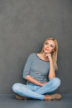 Pensive beauty. Beautiful young woman holding hand on chin and looking away with smile while sitting in lotus position against grey backgroundの写真素材