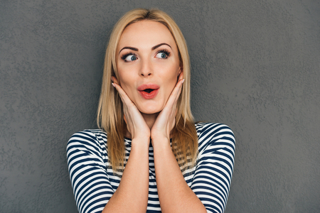 Oh! Beautiful young woman keeping hands on chin and looking surprised while standing against grey backgroundの写真素材