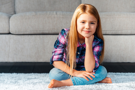 Maybe play? Cheerful little girl looking at camera with smile while sitting on the carpet in lotus position at homeの写真素材