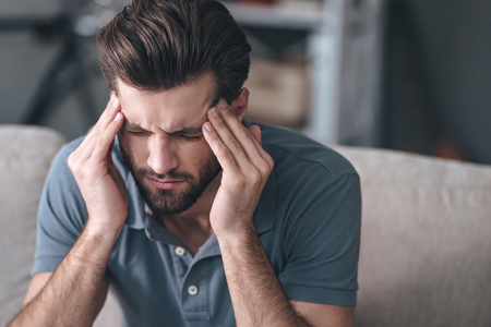 Feeling stressed. Frustrated handsome young man touching his head and keeping eyes closed while sitting on the couch at homeの写真素材