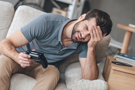 Lost this battle. Desperate young man holding joystick and keeping his hand on forehead while sitting on the couch at homeの写真素材