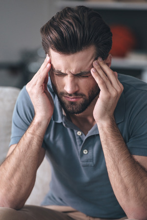 Feeling exhausted. Frustrated  young man touching his head and keeping eyes closedwhile sitting on the couch at homeの写真素材