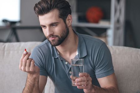 Just one pill can help. Handsome young man holding a glass of water and looking at a pill in his hand while sitting on the couch at homeの写真素材