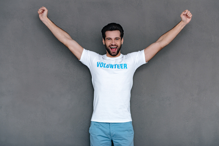 Hurray! Cheerful young man in volunteer t-shirt reaching out his armsand looking at camera with smile while standing against grey backgroundの写真素材