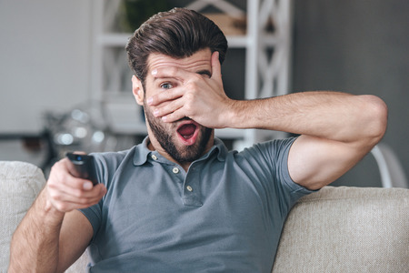 Horror movie. Handsome young man holding remote control and watching TV through his fingers while sitting on the couch at homeの写真素材