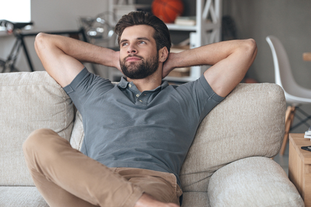 Time to gather all ideas in his head. Handsome young man keeping hands behind his head and looking thoughtful while sitting on the couch at homeの写真素材
