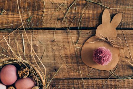 Ester Bunny. Top view of colored Easter eggs in bowl with hay and Easter bunny made from brown paper lying on wooden rustic tableの写真素材