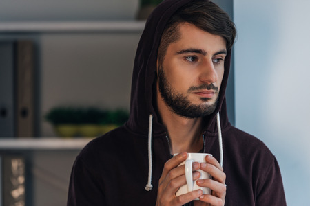Feeling frustrated. Portrait of pensive young man in hooded shirt holding coffee cup and looking away while standing at homeの写真素材