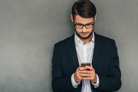 Business message. Confident young man in glasses using his smartphone while standing against grey backgroundの写真素材
