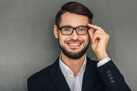 I clearly can see this! Portrait of cheerful young man looking at camera with smile and adjusting his glasses while standing against grey backgroundの写真素材