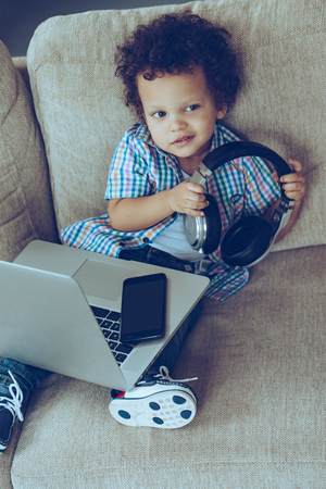 My favorite headphones! High angle view of little African baby boy holding headphones while sitting on the couch at home with laptop on his kneesの写真素材