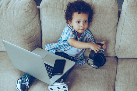 Can you bring my digital tablet please? High angle view of little African baby boy holding headphones and looking away while sitting on the couch at home with laptop and smartphone on his kneesの写真素材