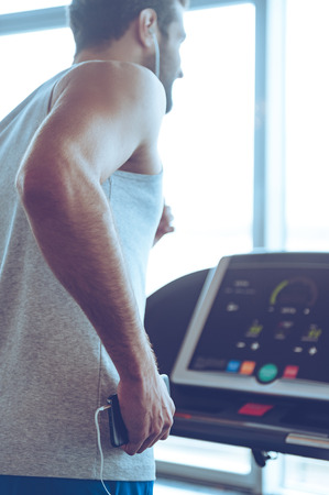 Exercising with great music. Rear view of young man in headphones running on treadmill in front of window at gym and holding his smart phone in his handの写真素材