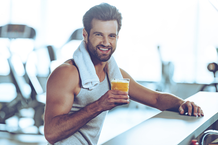 Sip of freshness after great workout. Side view of handsome young men in sportswear holding glass of fresh orange juice and looking at camera with smile while sitting at bar counter at gymの写真素材