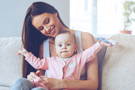 Here I am! Beautiful young woman holding baby girl with toy on her knee sand looking at her with...