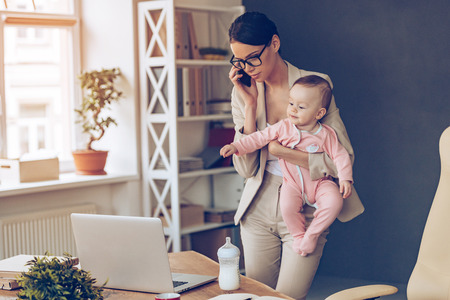 It is not easy to be a working mom! Young beautiful businesswoman talking on mobile phone and looking at laptop while standing with her baby girl at her working placeの写真素材