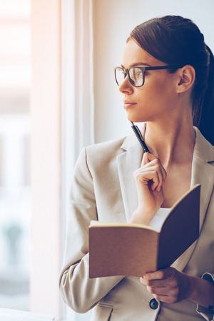 Finding the perfect solution. Close-up of pensive young beautiful businesswoman in glasses holding notebook and looking through window while standing at the windowsillの写真素材