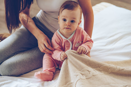 Time to start new day! Close-up part of little baby girl sitting on bed with her mother and pulling...